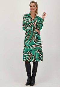 Groene jurk met gedurfde, abstracte strepen in het wit en bruin. Heeft een kraag, lange mouwen en een getailleerde taille, gecombineerd met zwarte kniehoge laarzen.