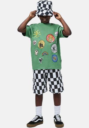 T-shirt di cotone verde con 10 toppe colorate, abbinata a pantaloncini a quadri bianchi e neri. Un cappello bucket a quadri bianco e nero completa il look.