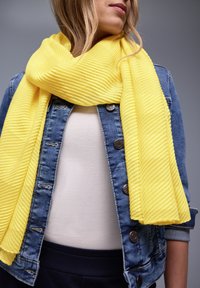 Femme portant une écharpe texturée jaune vif sur une chemise blanche et une veste en jean bleue, devant un fond gris uni.