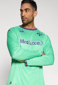 Zelen jopič z dolgimi rokavi z mrežastim teksturo, s vijoličnimi poudarki in logotipom "Mediacom" na sredini. Rokavi do komolcev in okrogel izrez na vratu.