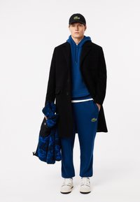 Cappotto nero su un felpa blu, pantaloni blu e sneakers bianche; tiene in mano una giacca blu con texture. Presenta un logo verde sui pantaloni.