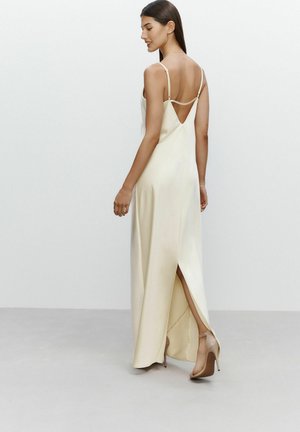 Abito maxi in satin color crema con spalline sottili, profonda scollatura a V sul retro e spacco laterale. Abbinato a scarpe con tacco nude. Texture liscia e colore uniforme.