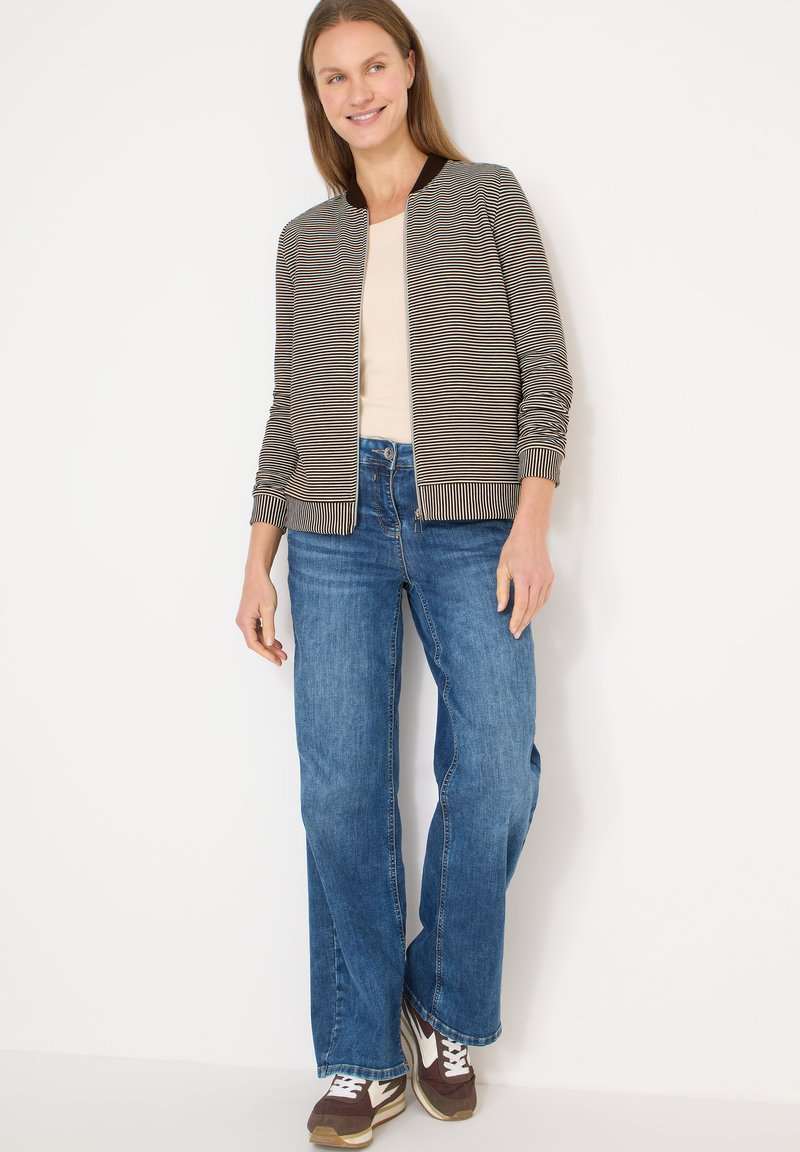 Veste bomber rayée avec un col sombre, portée sur un haut clair et un jean bleu à jambes larges ; associée à des baskets marron avec des accents blancs.