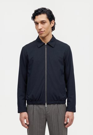 Un mannequin masculin porte une veste bleu marine zippée et un pantalon gris à fines rayures sur un fond blanc uni.