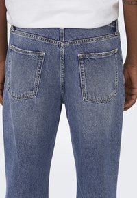 Jeans en denim bleu avec une coupe droite, deux poches arrière et des coutures visibles, présentant un léger délavage et des variations de texture.