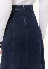 Dunkelblaue Jeansrock mit hoher Taille, Reißverschluss vorne und strukturiertem Design. Kombiniert mit einem weißen Langarmshirt, das oben sichtbar ist.