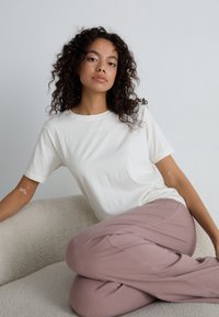 T-shirt en coton crème à manches courtes, arborant un branding subtil. Associé à un pantalon léger rose, posé de manière décontractée sur une surface texturée.