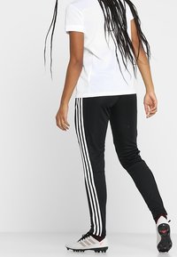 Black athletic pants with white vertical stripes, paired with a white polo shirt. A person is wearing cleats with a textured design.