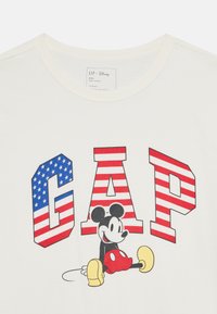 T-shirt en coton blanc avec un imprimé graphique de "GAP" en lettres rayées rouges et bleues, accompagné de Mickey Mouse portant un short rouge et des chaussures jaunes.