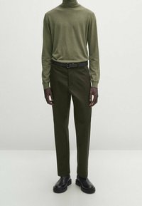 Olivengroen coltrui gecombineerd met donkergroene broek. Zwarte riem en zwarte enkellaarsjes. Effen texturen, slim fit ontwerp, neutrale achtergrond.
