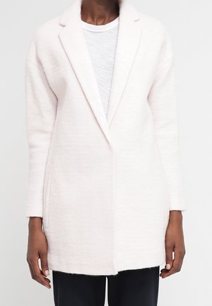 Femme portant un long manteau rose clair à col à revers par-dessus un haut blanc et un pantalon foncé, vue des épaules jusqu'au milieu des cuisses.