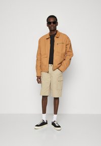 Tan bomuldsjakke med en lynlås lomme, sort t-shirt, beige cargo shorts og sorte sneakers med hvide såler, båret med mørke solbriller.