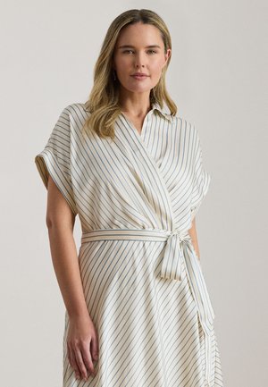 Naine, kes kannab kreemjat mähkekleit õhukeste siniste vertikaalsete triipudega ja vöökohal seotava vööga, seisab ühtlase tausta ees.