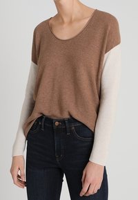 Brauner Pullover mit cremefarbenen Ärmeln, runder Ausschnitt, weiche Textur, entspannte Passform, kombiniert mit dunklen Jeans, die einen lässigen Look präsentieren.