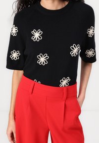 Pull en maille noire avec broderie de fleurs blanches, manches courtes, associé à un pantalon rouge vif et ajusté. Design simple et texturé.