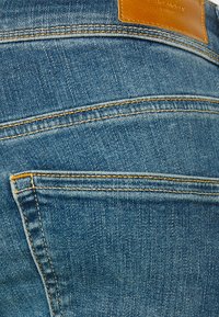 Denim kavbojke s klasičnim pet-pocket dizajnom, v srednji modri pranjeni barvi, subtilno teksturo in kontrastnim rumenim šivom. Usnjen logotip na pasu.