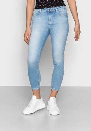 Femme portant un jean skinny bleu clair coupé au-dessus des chevilles et des baskets blanches à lacets, debout devant un fond clair uni.