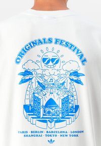 White T-shirt featuring a blue graphic design with a smiling sun, palm trees, speakers, and text "ORIGINALS FESTIVAL" with city names.