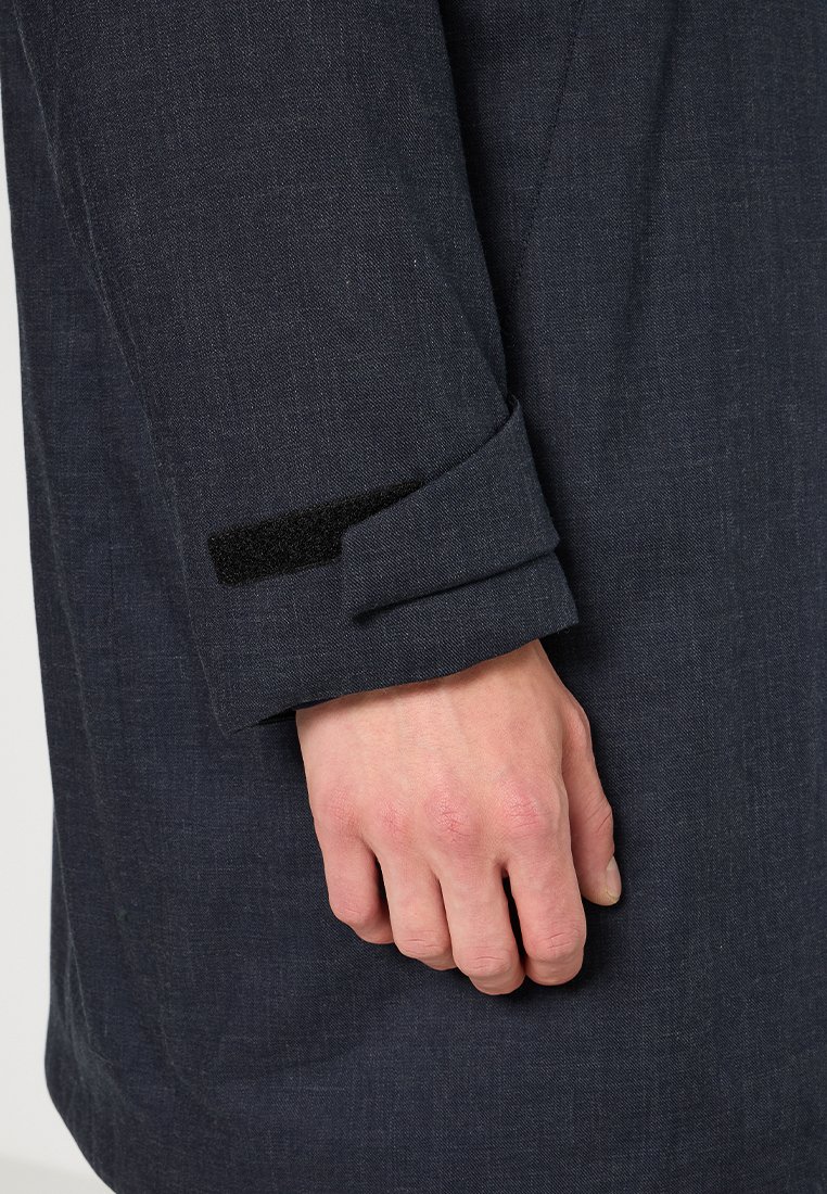 Manteau bleu foncé avec une finition texturée, présenté avec un poignet replié et un détail en velcro noir sur la manche. Main reposant sur le tissu.