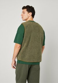Olive green sleeveless fleece vest with a textured, fluffy surface, trimmed in darker green, worn over a matching green shirt.