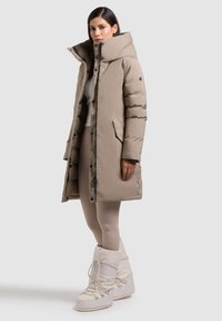 Bežs puffer mētelis ar augstu apkakli, ar spiedpogām, sānu kabatām un garu siluetu, kombinēts ar baltām sniega zābakiem.