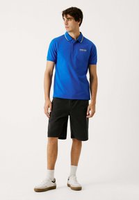 Niebieska koszulka polo z żółtymi akcentami na kołnierzyku, w połączeniu z czarnymi szortami, białymi trampkami i białymi skarpetkami. Casulowy, krótki krój rękawów.