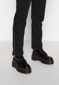 Stivali alla caviglia in pelle nera con suola spessa, design con lacci e dettagli di cucitura gialla, abbinati a jeans neri attillati con cerniere.