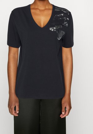 Persona con una camiseta negra de cuello en V y manga corta con diseños de lentejuelas negras en el hombro izquierdo y pantalones negros.