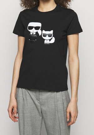 Černé tričko s bílými kreslenými postavičkami člověka a kočky s brýlemi na hrudi, nošené s šedými kalhotami.