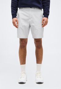 Άνδρας που φοράει ανοιχτό γκρι σορτς, λευκές κάλτσες, λευκά αθλητικά παπούτσια και σκουρόχρωμο μακρυμάνικο μπλουζάκι, στέκεται μπροστά σε απλό φωτεινό φόντο.