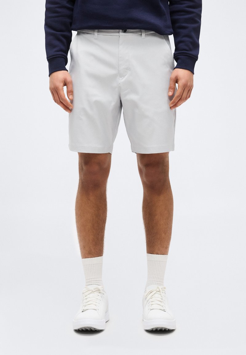 Άνδρας που φοράει ανοιχτό γκρι σορτς, λευκές κάλτσες, λευκά αθλητικά παπούτσια και σκουρόχρωμο μακρυμάνικο μπλουζάκι, στέκεται μπροστά σε απλό φωτεινό φόντο.