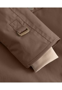 Manche de veste imperméable marron avec un doublure intérieure beige à revers. Comprend des accessoires en métal et des détails de couture renforcée. Texture lisse.
