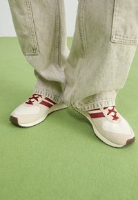Zapatillas deportivas beige con rayas y cordones rojos. Parte superior de material sintético texturizado y suela de goma. Combinadas con pantalones sueltos de color claro.