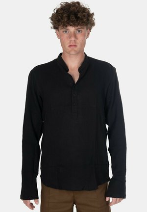Giovane uomo con capelli ricci indossa una camicia nera a maniche lunghe con bottoni e pantaloni marroni, in piedi contro uno sfondo chiaro e uniforme.