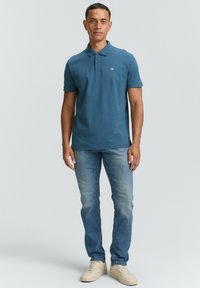 Camiseta polo en color azul verdoso con una textura sutil, tapeta de tres botones y logo en el pecho. Combinada con vaqueros de un azul claro y zapatillas beige.