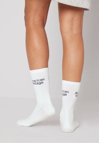 Calcetines de algodón blancos hasta el tobillo con textura acanalada y logo negro "American Vintage" impreso en la parte superior. Se muestran de lado sobre piernas desnudas.
