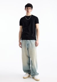 Tricou din bumbac negru, croială lejeră, asortat cu blugi ușor evazați de culoare bleu deschis. Pantofi sport albi cu tălpi bej și un colier din lanț argintiu ca accent.