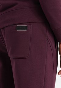 Burgunderfarbene Jogginghosen verfügen über eine Seitentasche mit einem rechteckigen schwarzen Logo-Patch, einer weichen Textur sowie einem gerippten Bund und Bündchen.