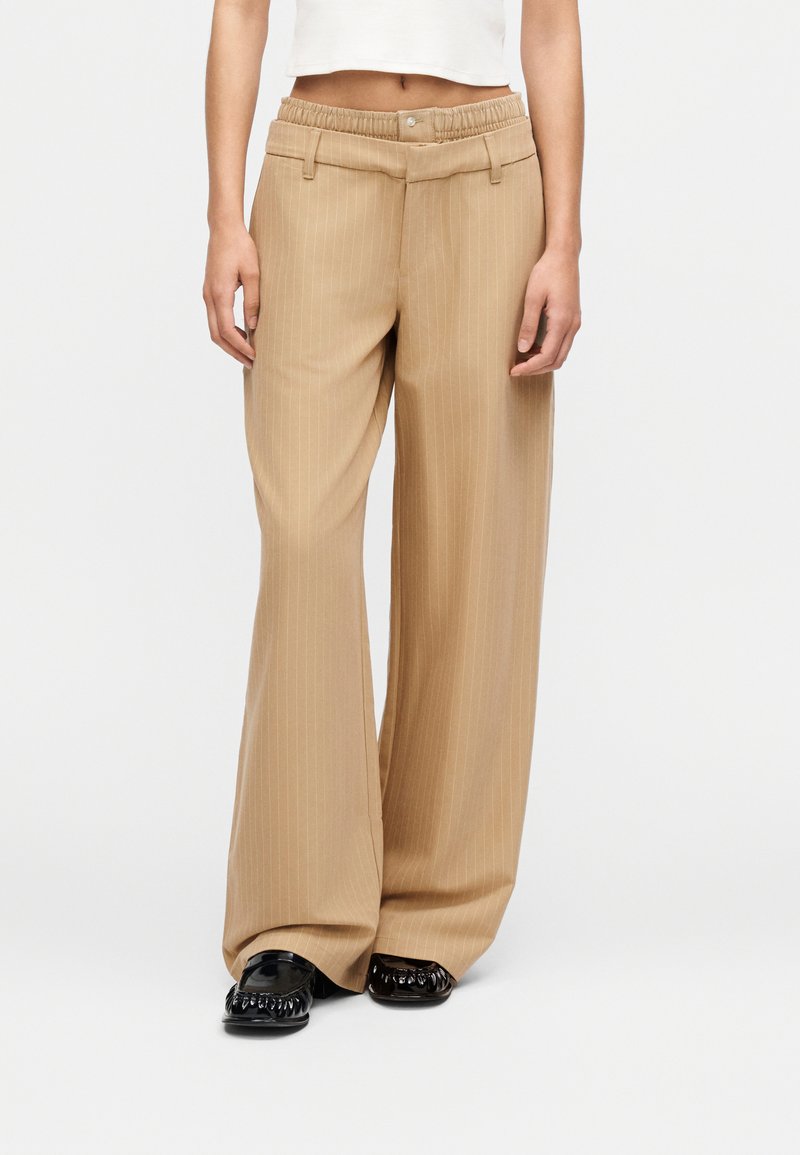 Άτομο που φοράει μπεζ παντελόνι με ρίγες και φαρδύ πόδι, μαύρα γυαλιστερά μοκασίνια και κοντό λευκό αμάνικο τοπ, στέκεται μπροστά σε λευκό φόντο.