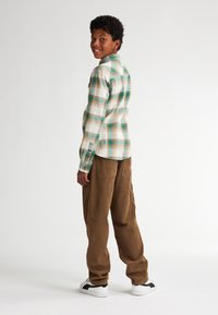 Kostkovaná košile s dlouhým rukávem v bílé barvě s zelenými a hnědými akcenty, spárovaná s hnědými manšestrovými kalhotami a bílými teniskami, pohled zezadu.