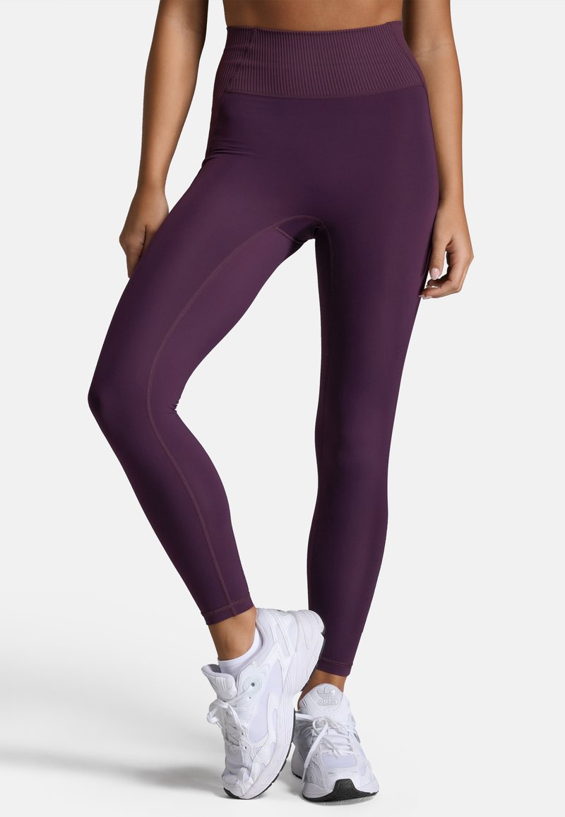 Paarse high-waisted legging met geribbelde tailleband, gladde textuur en contrasterende stiksels. Gekocht in combinatie met witte sportschoenen.