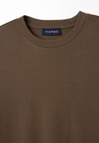T-shirt in cotone marrone con scollatura a girocollo. Presenta una texture morbida e un'etichetta con la scritta "PIOMBO" all'interno del colletto.