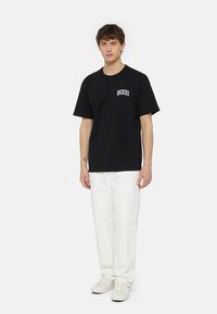Fekete pamut póló rövid ujjakkal és kerek nyakkal; a mellkasán kék "Dickies" logó hímezve. Fehér nadrággal viselve.