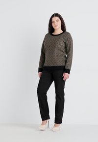 Pull en tricot avec un motif géométrique en or et noir, associé à un pantalon noir. Le modèle porte des talons beiges. Fond simple.
