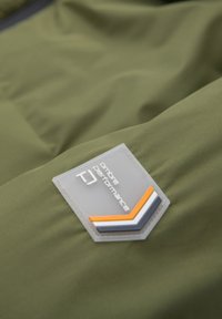 Tissu vert avec un écusson cousu portant l'inscription "Ombre Performance" et un motif chevron rayé en orange, blanc et gris.