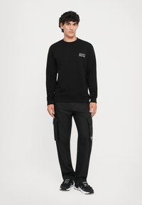 Sweatshirt preta com o logótipo "VERSACE COUTURE", combinado com calças cargo pretas com bolsos e um acabamento texturizado. Ténis pretos completam o look.