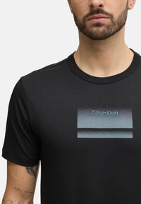 Camiseta negra hecha de una tela suave, con un gráfico rectangular en azul que incluye el texto "Calvin Klein" en una fuente moderna en la parte frontal.