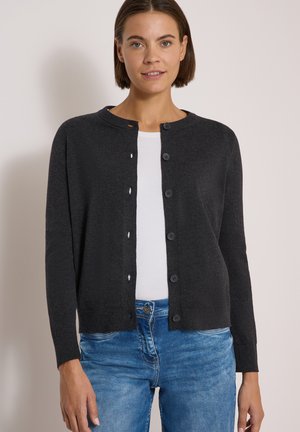 Femme portant un cardigan noir boutonné par-dessus un haut blanc et un jean bleu, debout devant un fond clair uni.