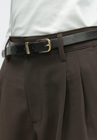 Black leather belt featuring a gold metal buckle, worn over brown pleated trousers. The fabric is smooth with a subtle sheen.
