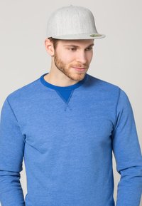 Grijze pet met een vlakke rand, gestructureerde stof en een klein logo. Draagt bij een blauwe sweatshirt met een ronde hals en geribde boorden.